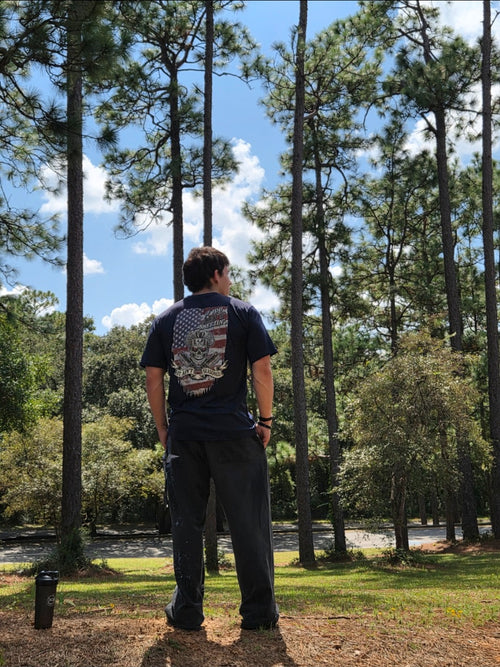 Back view of the navy Seasoned American T-Shirt worn outdoors among pine trees, showing the American flag and chef-skull artwork.